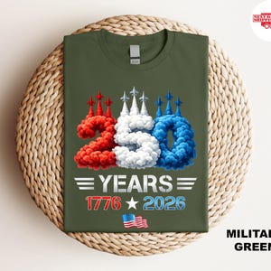 Puede incluir: Camiseta verde militar con un dise&ntilde;o gr&aacute;fico que celebra 250 a&ntilde;os. El dise&ntilde;o incluye el n&uacute;mero "250" en humo rojo, blanco y azul, el texto "YEARS", las fechas "1776" y "2026", y una bandera estadounidense.