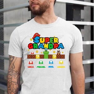 Op de afbeelding: Wit T-shirt met een Super Opa-afbeelding in Mario-stijl. De afbeelding bevat een groene pijp, munten en vier paddenstoelfiguren met de namen Luca, Michael, Noel en Lucia.