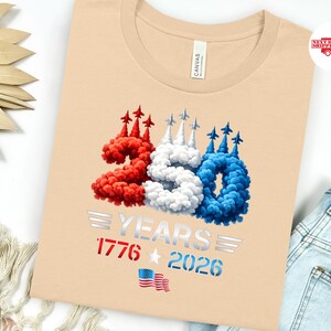 Puede incluir: Una camiseta beige con un dise&ntilde;o patri&oacute;tico que celebra 250 a&ntilde;os. El n&uacute;mero "250" est&aacute; en rojo, blanco y azul, imitando estelas de humo con peque&ntilde;os aviones. El texto "YEARS 1776 2026" est&aacute; debajo, junto con una peque&ntilde;a bandera estadounidense.