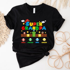 Op de afbeelding: Zwart T-shirt met een kleurrijke afbeelding van een Super Mario Bros. level. De tekst "SUPER GRANDPA" staat bovenaan de afbeelding. De namen "LUCA", "MICHAEL", "NOEL" en "LUCIA" staan onderaan de afbeelding.