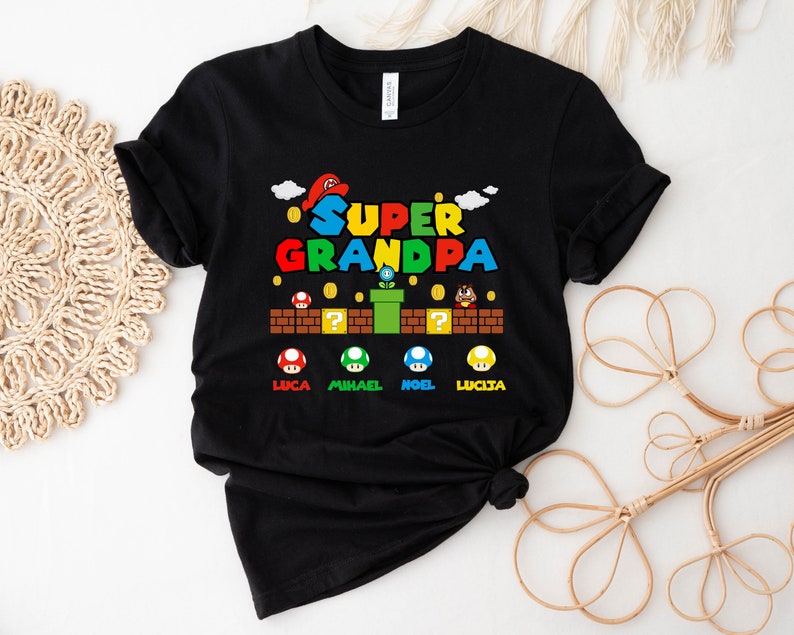 Op de afbeelding: Zwart T-shirt met een kleurrijke afbeelding van een Super Mario Bros. level. De tekst "SUPER GRANDPA" staat bovenaan de afbeelding. De namen "LUCA", "MICHAEL", "NOEL" en "LUCIA" staan onderaan de afbeelding.