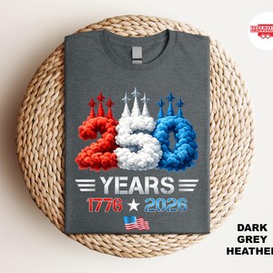 Puede incluir: Camiseta gris oscuro jaspeado con un dise&ntilde;o patri&oacute;tico. El dise&ntilde;o presenta el n&uacute;mero "250" en rojo, blanco y azul, con el texto "YEARS 1776 2026" y una peque&ntilde;a bandera estadounidense. La camiseta se muestra sobre una superficie tejida.