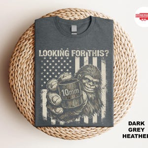 May include: Dark grey heather t-shirt featuring a graphic of a smiling Bigfoot holding a 10mm object in its hand, set against a distressed American flag design. The text "LOOKING FOR THIS?" is above the graphic.