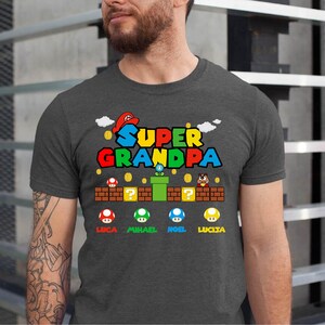 Op de afbeelding: Een donker grijs gem&ecirc;leerd t-shirt met een Super Mario Bros.-thema. De afbeelding bevat de tekst "Super Grandpa" met een paddenstoel, pijp en vraagtekenblok. De namen Luca, Mikael, Noel en Lucia staan onder de afbeelding.