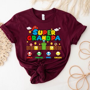 Op de afbeelding: Bordeauxkleurig T-shirt met een Super Opa-afbeelding met een Mario-thema met de namen Luca, Michael, Noel en Lucia.