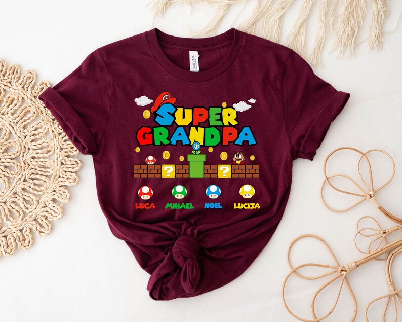 Op de afbeelding: Bordeauxkleurig T-shirt met een Super Opa-afbeelding met een Mario-thema met de namen Luca, Michael, Noel en Lucia.
