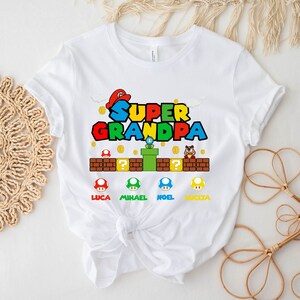 Op de afbeelding: Wit T-shirt met een Super Mario Bros.-thema met de tekst "Super Opa" in rood, groen, blauw en geel. De afbeelding bevat een paddenstoel, een pijp en een vraagtekenblok. Er zijn vier kleinere paddenstoelen met de namen Luca, Michael, Noel en Lucia.