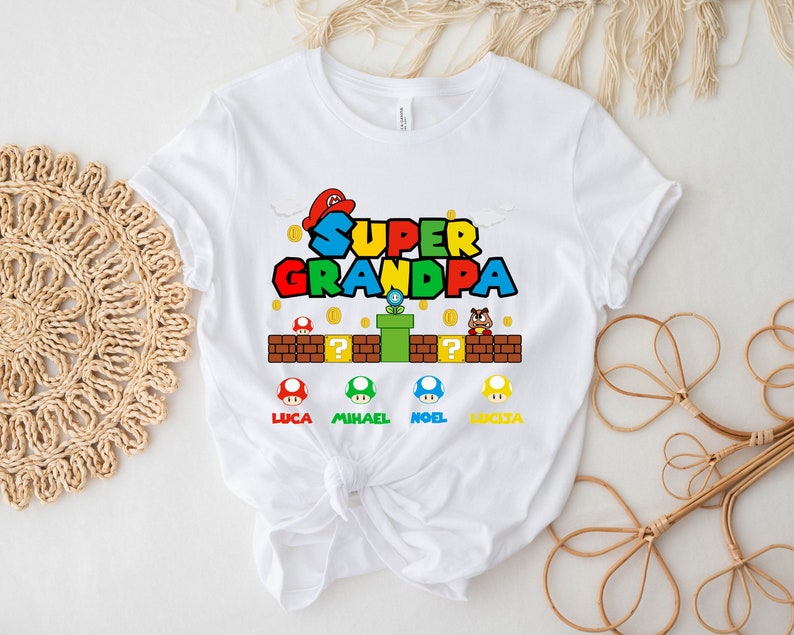 Op de afbeelding: Wit T-shirt met een Super Mario Bros.-thema met de tekst "Super Opa" in rood, groen, blauw en geel. De afbeelding bevat een paddenstoel, een pijp en een vraagtekenblok. Er zijn vier kleinere paddenstoelen met de namen Luca, Michael, Noel en Lucia.