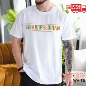 May include: White t-shirt with the text "The Grandpalorian The Best Grandpa in the Galaxy" in colorful letters.
