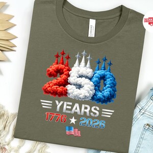 Puede incluir: Camiseta verde oliva con un dise&ntilde;o patri&oacute;tico. El dise&ntilde;o presenta el n&uacute;mero 250 en rojo, blanco y azul, con el texto "YEARS 1776 2026" y una bandera americana. La camiseta est&aacute; hecha de un material suave.