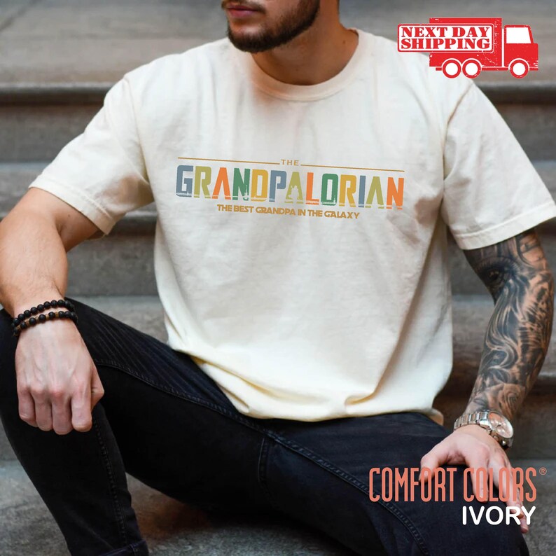 May include: A white t-shirt with the text "THE GRANDPALORIAN THE BEST GRANDPA IN THE GALAXY" in a retro font with colorful letters. The shirt is being worn by a person with tattoos on their arm.