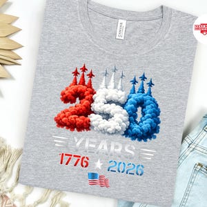 Puede incluir: Una camiseta gris con un dise&ntilde;o patri&oacute;tico. El dise&ntilde;o presenta los n&uacute;meros "250" en rojo, blanco y azul, con el texto "YEARS 1776 2026" y una bandera estadounidense. La camiseta est&aacute; hecha de un material suave.