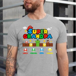Op de afbeelding: Een grijs T-shirt met de tekst "Super Opa" in een kleurrijk videospel-lettertype. Het shirt heeft een cartoonfiguur met een rode hoed en een groene pijp. Onder de tekst staan vier paddenstoelfiguren met de namen Luca, Michael, Noel en Lucca.