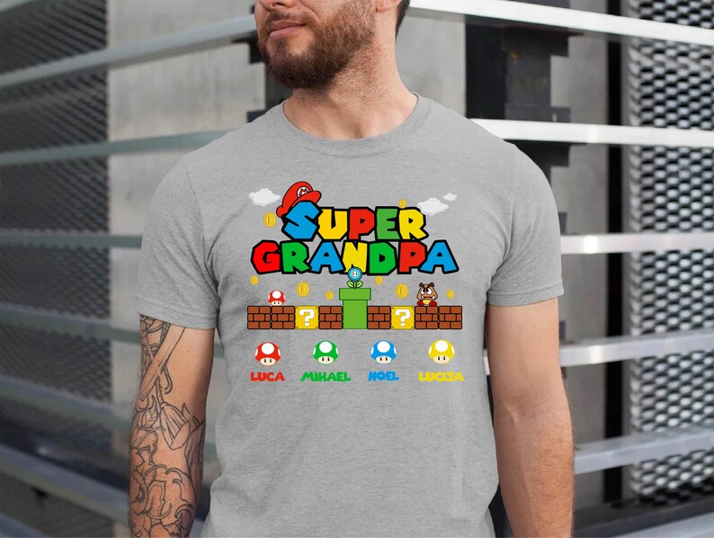 Op de afbeelding: Een grijs T-shirt met de tekst "Super Opa" in een kleurrijk videospel-lettertype. Het shirt heeft een cartoonfiguur met een rode hoed en een groene pijp. Onder de tekst staan vier paddenstoelfiguren met de namen Luca, Michael, Noel en Lucca.