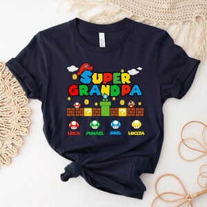 Op de afbeelding: Een donkerblauw T-shirt met een Super Opa-afbeelding. De afbeelding is in Super Mario Bros.-stijl met een paddenstoel- en pijpdesign. De namen Luca, Michael, Noel en Lucia staan onder de afbeelding gedrukt.