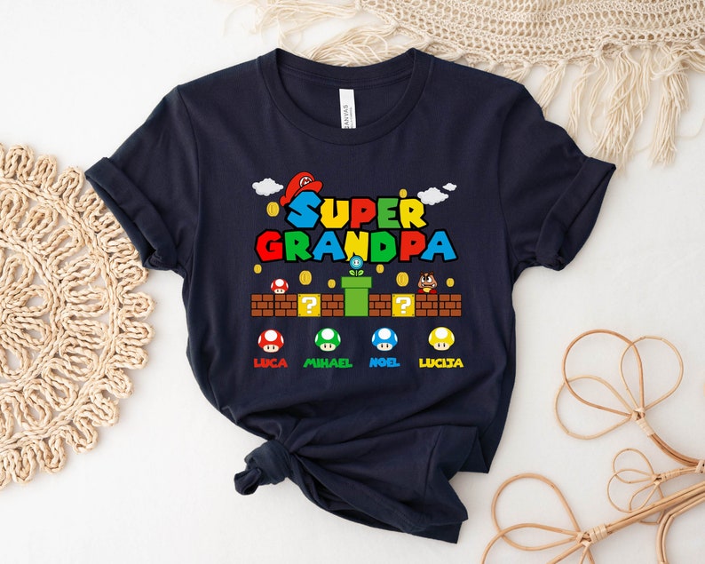 Op de afbeelding: Een donkerblauw T-shirt met een Super Opa-afbeelding. De afbeelding is in Super Mario Bros.-stijl met een paddenstoel- en pijpdesign. De namen Luca, Michael, Noel en Lucia staan onder de afbeelding gedrukt.