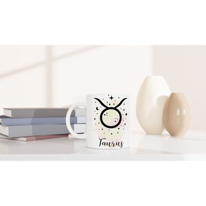 May include: White ceramic mug with a black and white illustration of the Taurus zodiac sign surrounded by stars. The mug has a watercolor-like background with shades of pink, yellow, and orange. The word "Taurus" is printed in black below the illustration.