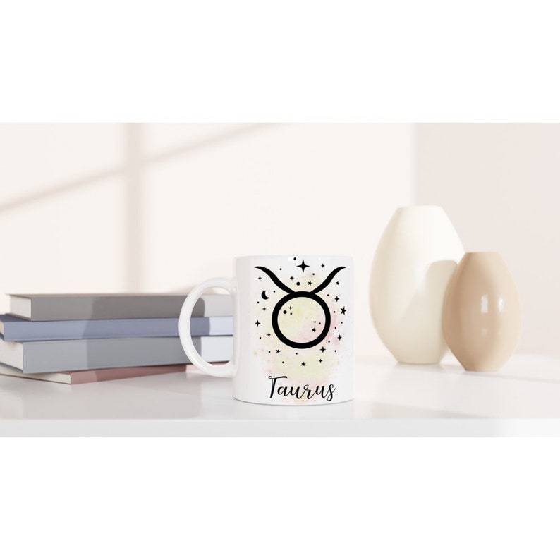 May include: White ceramic mug with a black and white illustration of the Taurus zodiac sign surrounded by stars. The mug has a watercolor-like background with shades of pink, yellow, and orange. The word "Taurus" is printed in black below the illustration.