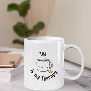 Puede incluir: Taza de cerámica blanca con un diseño en negro y amarillo. La taza presenta una taza de té de dibujos animados con una cara sonriente y el texto "Tea is my Therapy".