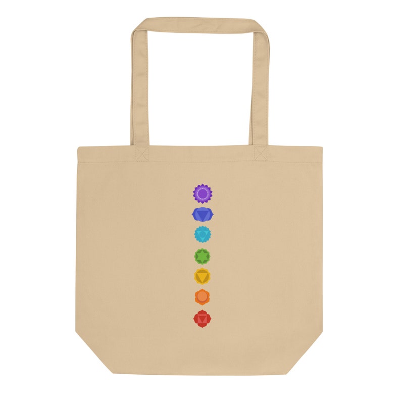 Puede incluir: Una bolsa de tela beige con una fila vertical de siete s&iacute;mbolos de chakras coloridos impresos en la parte delantera.