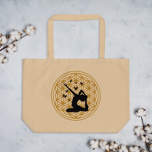 Puede incluir: Una bolsa de tela beige con un diseño circular dorado que presenta una silueta de una persona en una postura de yoga con mariposas volando a su alrededor. El diseño circular es un patrón de flor de la vida.
