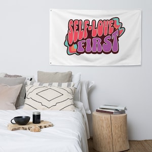 Può includere: Un arazzo bianco con il testo "SELF-LOVE FIRST" in un font retrò e colorato.