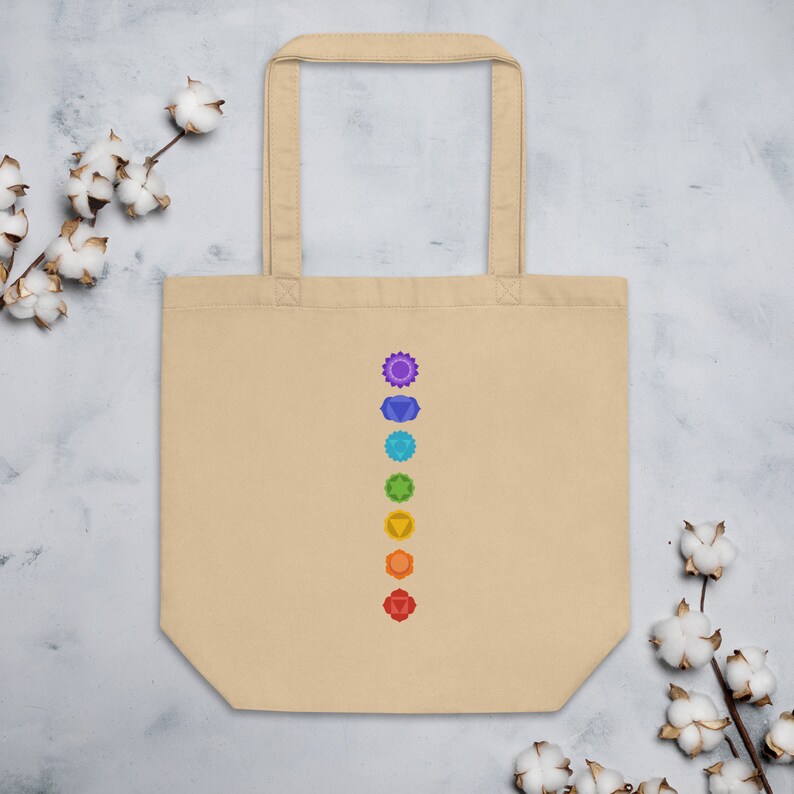 Puede incluir: Una bolsa de tela beige con una fila vertical de siete s&iacute;mbolos de chakras coloridos impresos en la parte delantera. Los s&iacute;mbolos est&aacute;n en los colores de los chakras: rojo, naranja, amarillo, verde, azul, &iacute;ndigo y violeta.