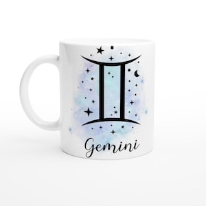 Puede incluir: Taza de cerámica blanca con un fondo de estilo acuarela en degradado azul y morado. La taza tiene un símbolo del zodíaco Géminis negro con estrellas y una luna creciente. La palabra "Géminis" está escrita en cursiva negra debajo del símbolo.
