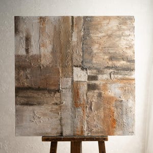 May include: Abstract painting on canvas with a textured, layered look in shades of brown, beige, and white. The painting is displayed on a wooden easel.