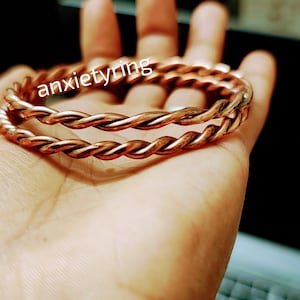 May include: Two copper bracelets, twisted together, in a hand. The bracelets are a reddish-brown color and have a shiny finish. The text "anxietyring" is visible on the bracelets.
