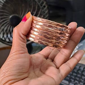 Puede incluir: Una pila de brazaletes de cobre martillado. Cada brazalete tiene una superficie texturizada y un color marrón rojizo cálido. Los brazaletes se sostienen en una mano, mostrando su diseño y artesanía. El texto "anxietyring" es visible.