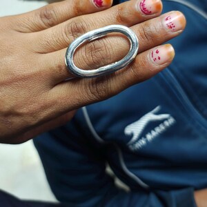 Peut inclure: Une bague ovale en argent avec une surface lisse et arrondie. La bague est portée sur le majeur d'une main avec des ongles peints.