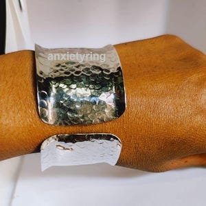 Op de afbeelding: Een zilveren gehammerde armband met een getextureerd oppervlak. De armband is open en heeft een licht gebogen vorm.