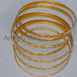 Könnte beinhalten: Ein Set aus sechs goldfarbenen, gehämmerten Metallarmreifen. Die Armreifen sind übereinander gestapelt und erzeugen so einen geschichteten Look.