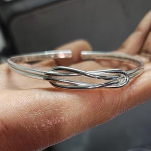May include: A silver knot bangle bracelet. The bracelet is made of two strands of silver that are twisted together to form a knot.