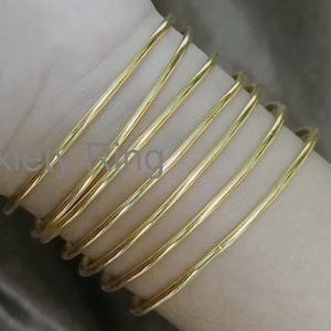 Puede incluir: Un conjunto de ocho pulseras de brazalete doradas apiladas en el brazo de una persona. Las pulseras son todas lisas y brillantes.