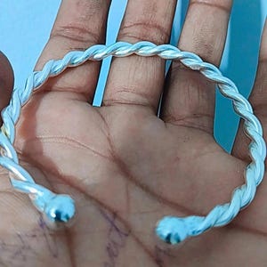 May include: A silver-coloured, twisted-rope design bracelet. The bracelet has a smooth, polished finish and is open-ended with rounded tips. The bracelet is a cuff style and appears to be made of metal.