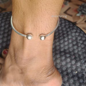 Könnte beinhalten: Ein silberfarbenes Fußkettchen mit schlichtem, offenem Design. Die Enden des Fußkettchens haben kleine, runde Kugelakzente. Der Text "anxietyring" ist im Hintergrund sichtbar.