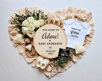 Op de afbeelding: Een houten bord met de tekst "One more to adore! Baby Anderson #3 August 2025" omringd door witte rozen, een baby-romper met de tekst "Here we grow again!" en andere baby-gerelateerde items.