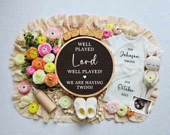 May include: A gender reveal announcement with a black and white sign that reads "Well Played Lord Well Played! We are having twins!"  The sign is surrounded by pink and yellow flowers, two white onesies, one with the text "The Johnson Twins" and the other with the text "Due October 2025", a crocheted bunny, and a wooden block spelling "TWINS".