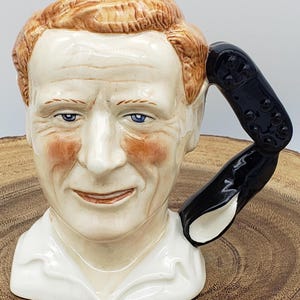 May include: A ceramic mug shaped like a man's head, featuring red hair, blue eyes, and a white shirt. The handle is a black, curved shape. The mug is set on a wooden surface.