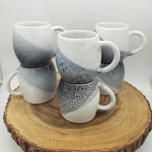 Könnte beinhalten: Sechs Keramikbecher mit einem einzigartigen Design. Jeder Becher hat einen weißen Boden, ein diagonales Band aus gesprenkeltem Grau und einen weißen Henkel. Die Becher sind auf einer Holzscheibe gestapelt, was ihre handwerkliche Optik unterstreicht.