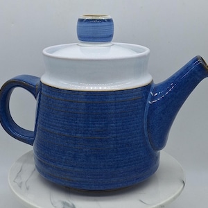 Puede incluir: Una tetera de cerámica con un cuerpo de color azul intenso con líneas horizontales grabadas y una tapa blanca en contraste. La tetera tiene un asa curva en el lado izquierdo y un pico cónico en el lado derecho, con un pomo redondeado en la tapa.