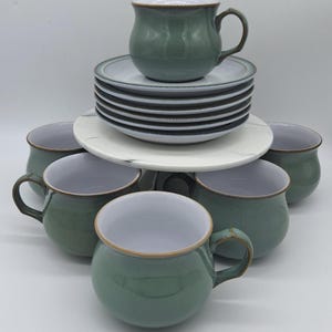 May include: A set of six sage green ceramic cups with white interiors and brown rims, accompanied by a stack of six matching saucers. The cups feature a rounded, bulbous shape with a single handle, while the saucers have a flat, circular design.