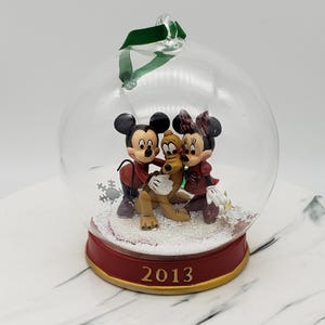 Puede incluir: Una bola de Navidad de cristal transparente con figuras de Mickey Mouse, Minnie Mouse y Pluto. El adorno tiene una base roja con el año "2013" en letras doradas. Una cinta verde está unida para colgar.