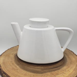 Può includere: Una teiera in ceramica bianca con coperchio e manico. La teiera è appoggiata su una superficie di legno. La teiera ha un beccuccio e un manico. La teiera è di colore bianco pieno.