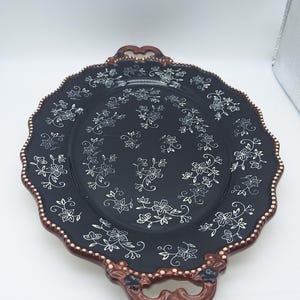 Könnte beinhalten: Ein ovales Serviertablett aus Keramik mit glänzend schwarzer Oberfläche und weißem Blumenmuster. Das Tablett hat einen gewellten Rand mit brauner Umrandung und kleinen weißen Punkten sowie zwei integrierte Griffe an den Enden mit braunen Details.