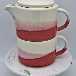 May include: A ceramic teapot and matching cup stacked vertically, featuring a two-tone glaze with a creamy off-white top section and a deep rose-pink bottom section. The teapot has a lid, a side handle, and a spout, while the cup has a side handle.