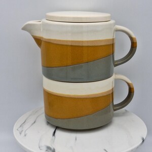 May include: A stackable ceramic teapot and mug set featuring a horizontal colour-blocked design in cream, mustard yellow, and slate grey. The teapot has a lid and a side handle, while the mug features a matching handle and curved silhouette.