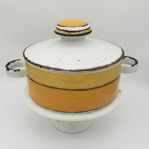 Op de afbeelding: Een keramische kookpot met deksel, in wit, geel en oranje. De pot heeft twee handvatten en staat op een witte sokkel. Het deksel heeft een afgeronde knop met zwarte en witte strepen. Geschikt om te koken.
